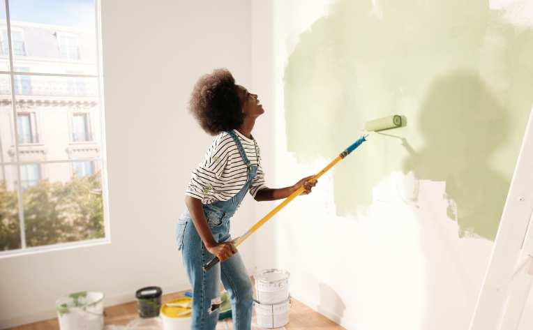 woman using paint roller to paint over white wall in apartment with green paint